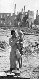 Frau mit Kind auf dem Arm vor Trümmern